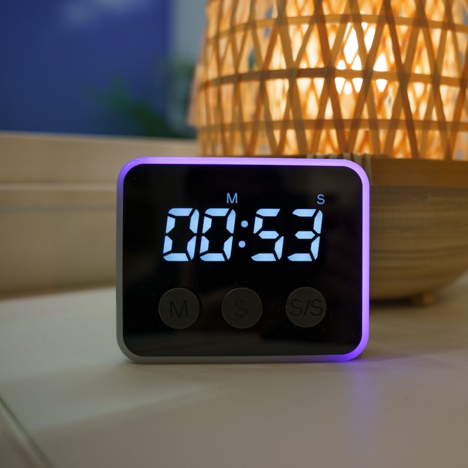 Digital timer with a purple border on a surface, blurred lamp in the background