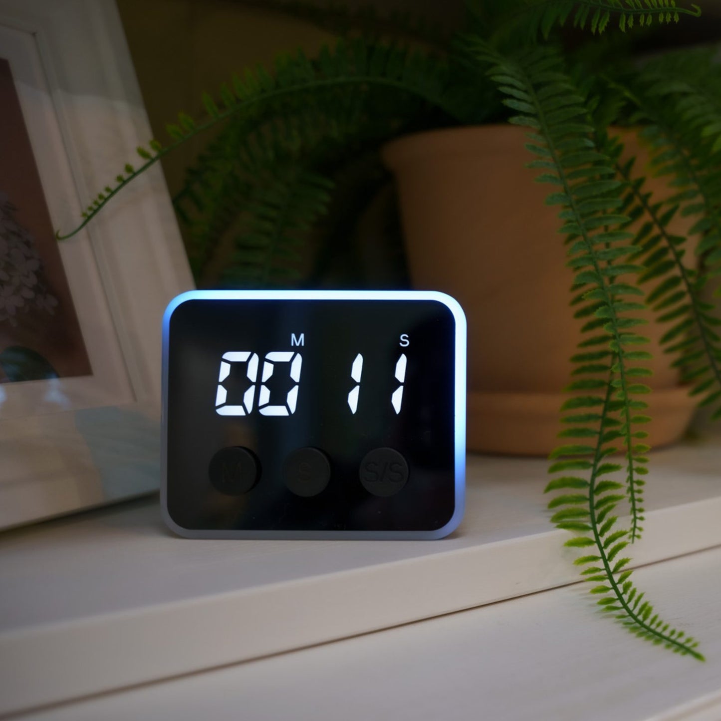 Looptimer visual timer with blue LED light reading 11 seconds on a white shelf next to a fern and framed picture